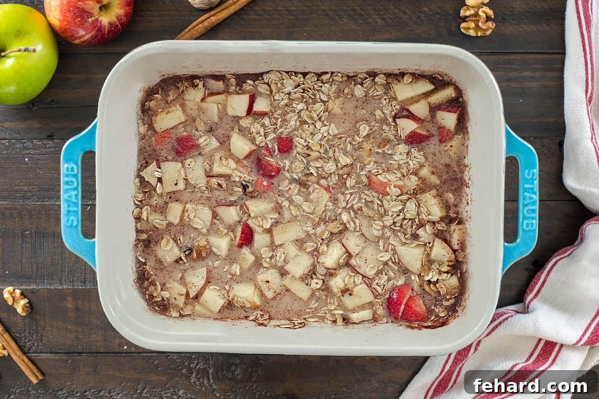 Oats, diced apples, walnuts, milk, spices, and an egg are thoroughly mixed together in a blue baking dish, surrounded by fresh apples and cinnamon sticks.