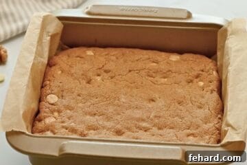 Freshly baked blondies in a metal pan lined with parchment paper, displaying golden-brown edges and a tender center.