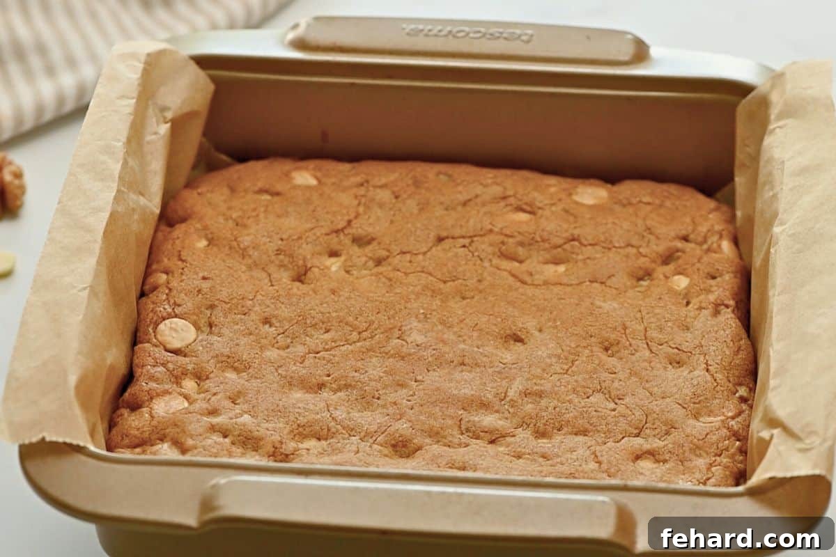 Freshly baked blondies, displaying golden-brown edges and a perfectly set yet tender center, still warm in the metal baking pan lined with parchment paper, fresh from the oven.