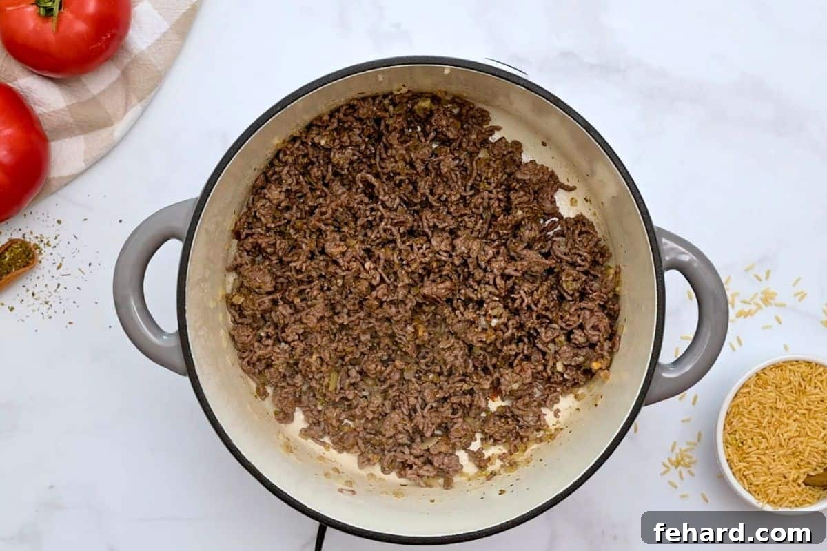 Healthy Stuffed Pepper Casserole Prep Ground beef browned with diced onion, minced garlic, and dried seasonings in a large stock pan, with uncooked brown rice visible next to it.