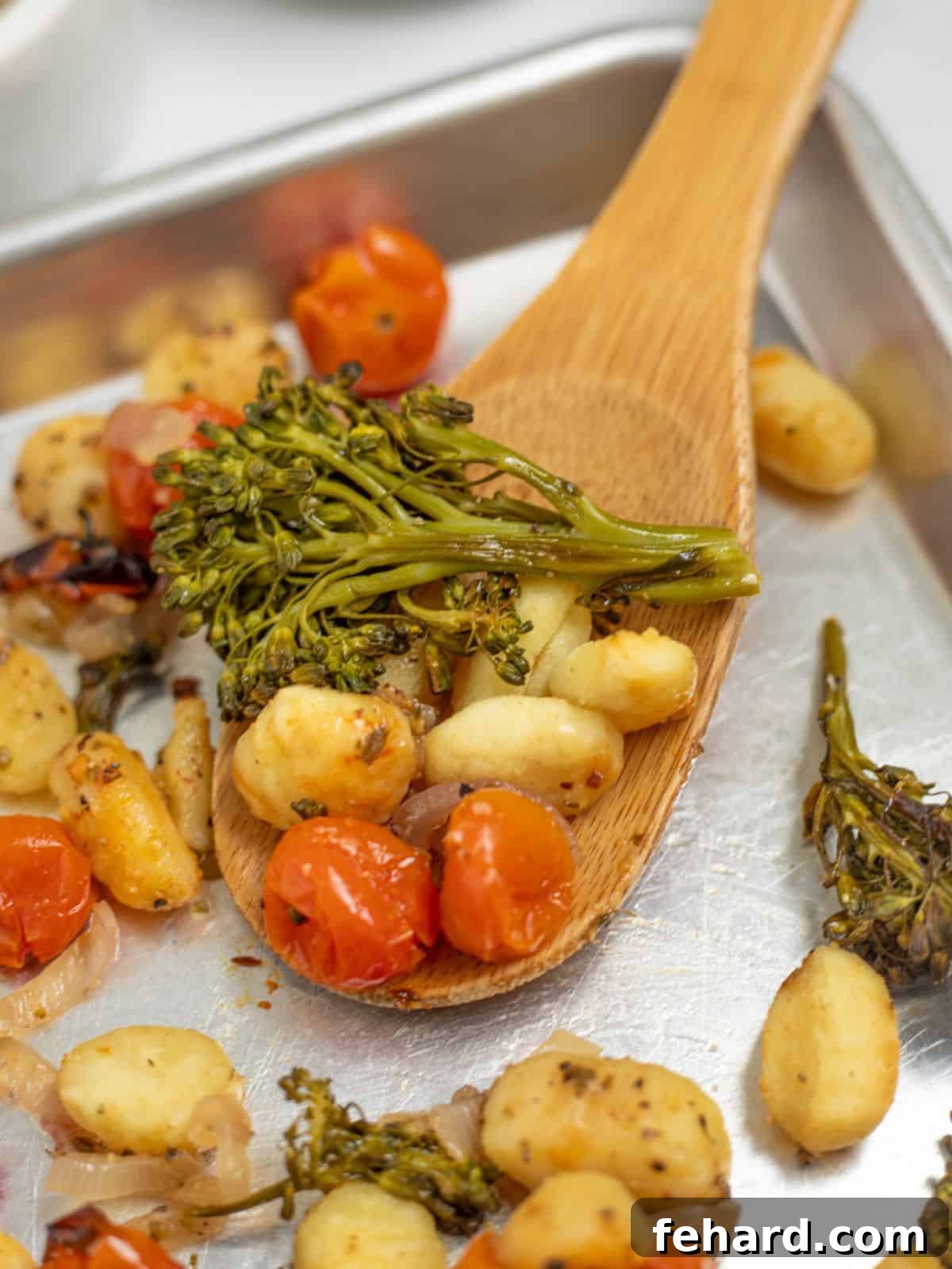 Wooden spoon scooping up roasted sheet pan gnocchi, tomatoes, and broccolini seasoned with Italian seasoning, highlighting its golden crisp texture.