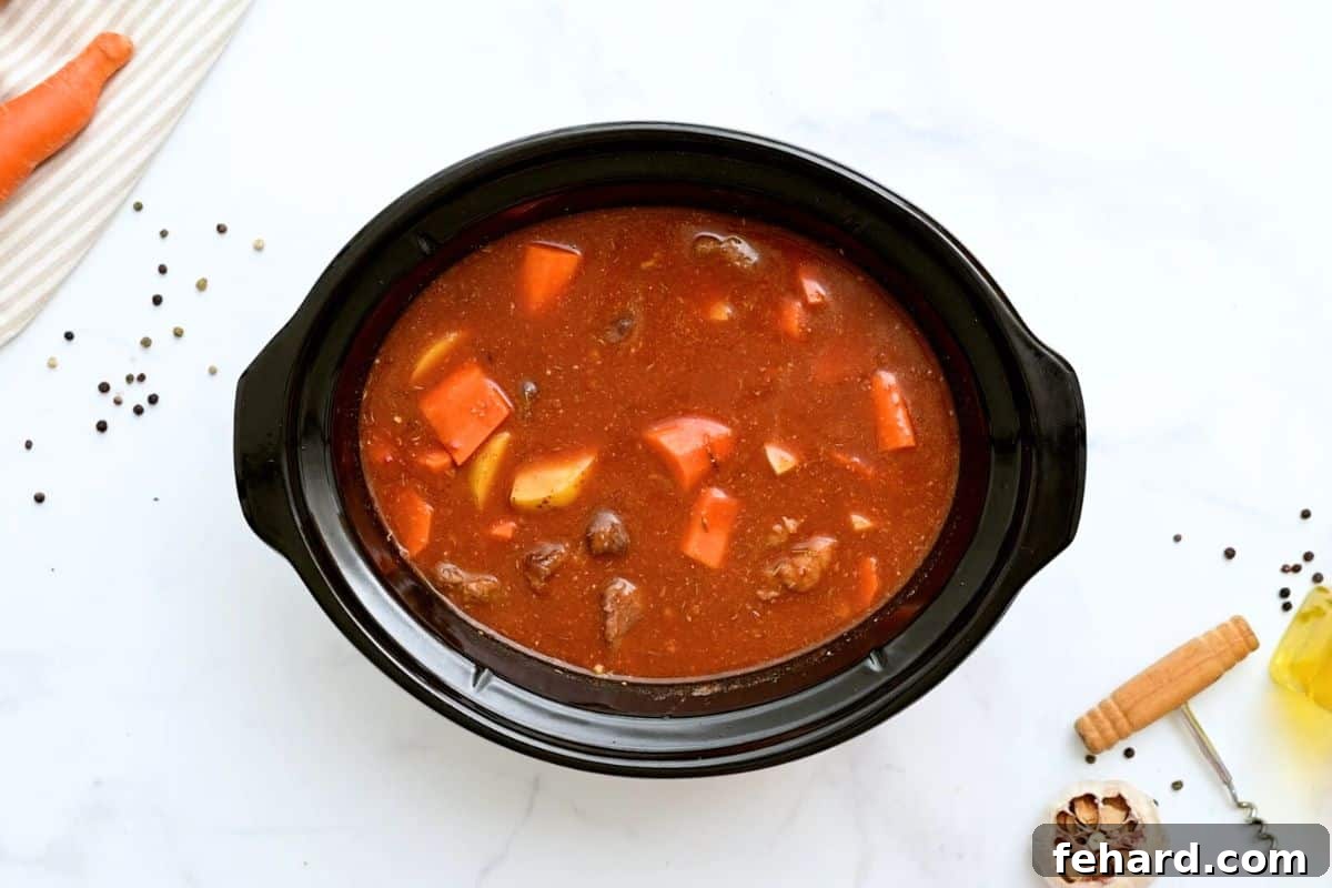 All the core ingredients for beef stew – browned beef, rich beef stock, tomato paste, potatoes, and carrots – neatly combined in the slow cooker, ready for simmering.