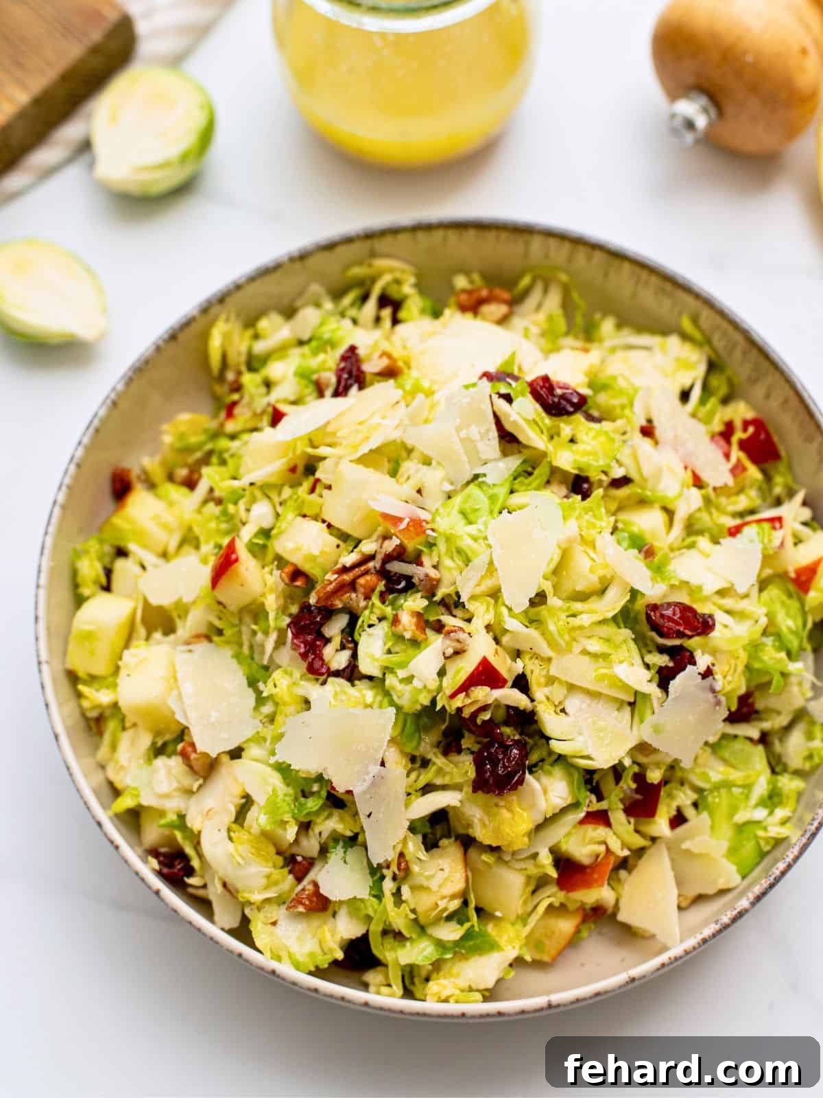 A vibrant Shaved Brussels Sprouts Salad in a rustic white serving bowl, glistening with a honey lemon dressing, and adorned with bright red diced apples, plump dried cranberries, delicate shaved Parmesan cheese, and golden chopped toasted pecans. A small decorative bottle of the dressing is visible blurred in the background.