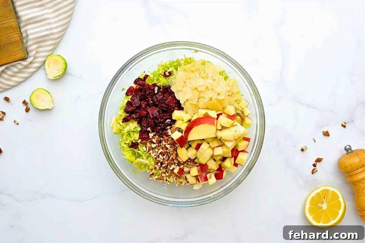 A large mixing bowl filled with marinated shaved Brussels sprouts, now beautifully topped with colorful diced apples, vibrant dried cranberries, delicate shaved Parmesan cheese, and crunchy chopped toasted pecans, eagerly awaiting a final toss.