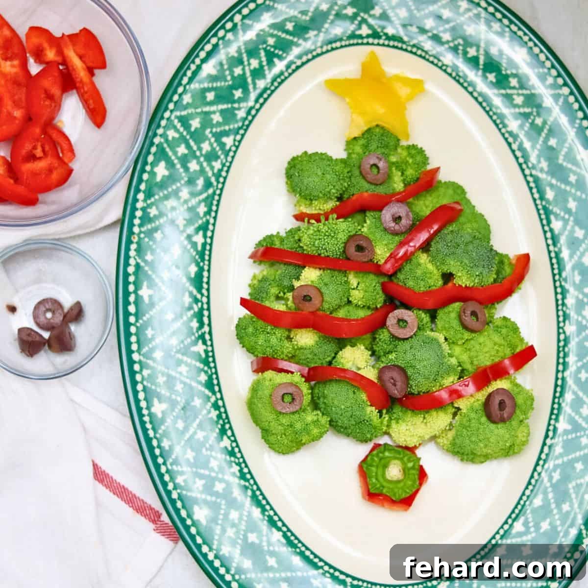 A festive Christmas tree made with fresh broccoli florets, vibrant red peppers, and black olive ornaments, arranged on a white platter with dip.