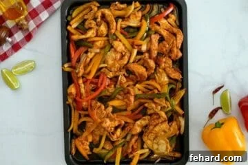 Sliced chicken breasts, sliced bell peppers, and sliced onions tossed with seasonings on rimmed sheet pan after baking.