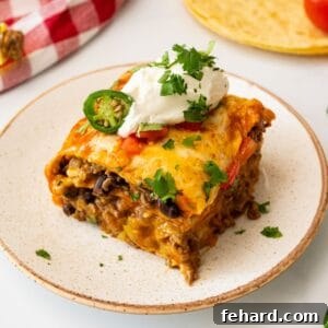 A slice of stacked beef enchilada casserole, revealing layers of ground beef, beans, cheese, and tortillas, topped with a dollop of sour cream and fresh cilantro.