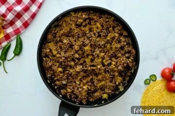 The cooked ground beef mixture combined with enchilada sauce, green chiles, and beans in a skillet.