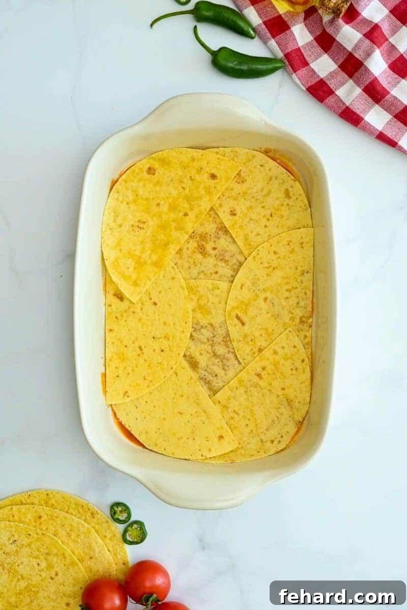 A 9x13-inch baking dish showing the first layer of the casserole: enchilada sauce topped with a layer of corn tortillas.