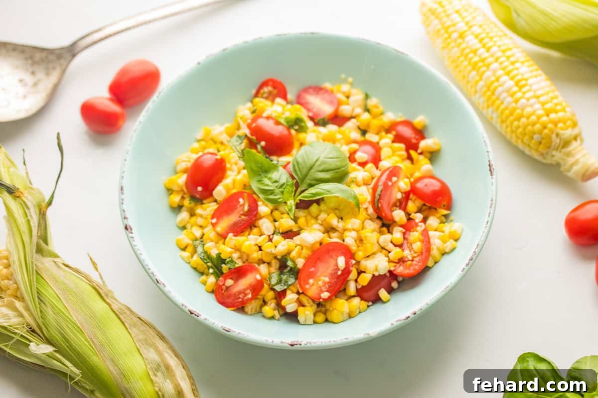 Fresh corn and tomato salad with basil in blue bowl with corn on the cob and grape tomatoes on the counter.