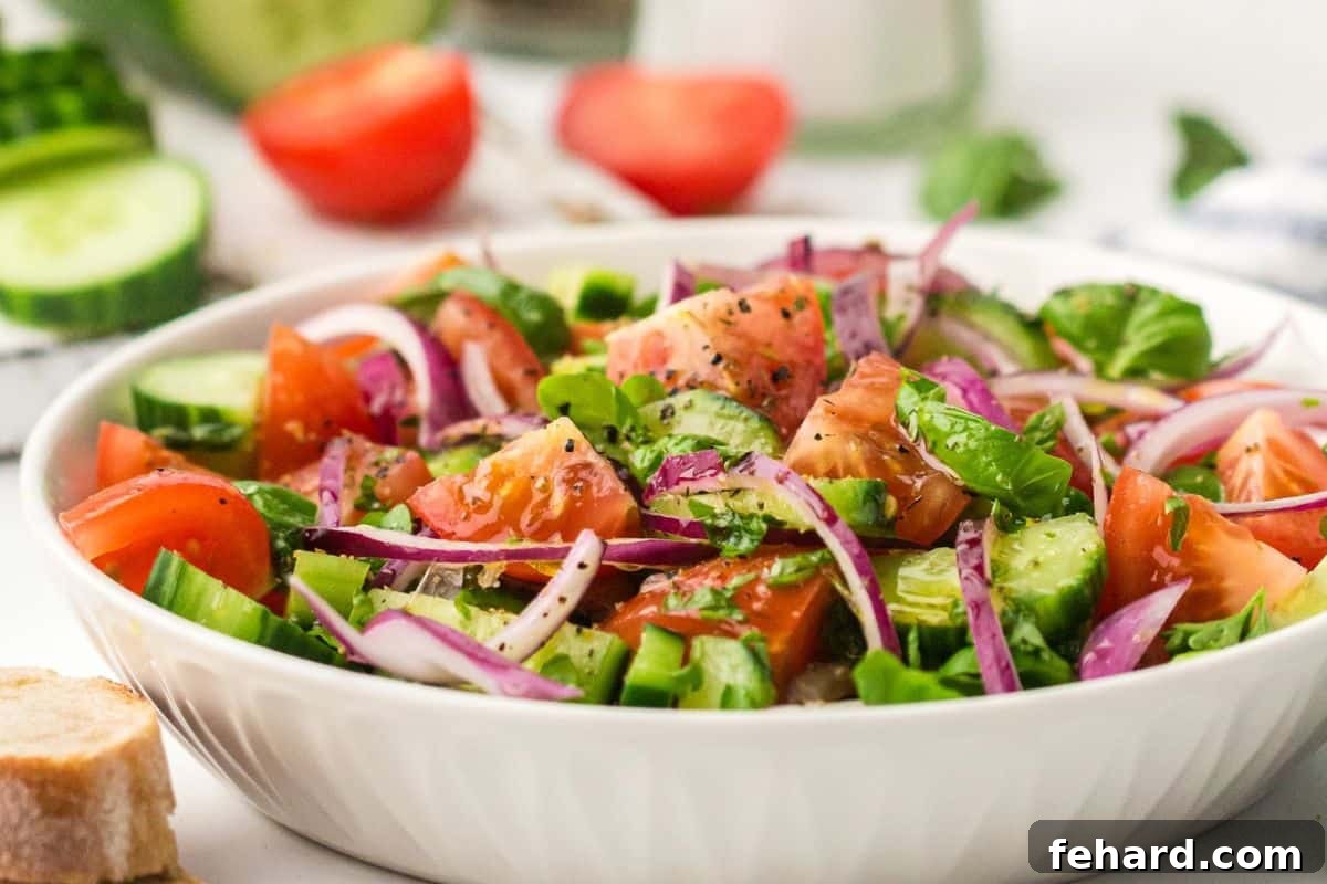 Cucumber, tomato, and red onion salad in white bowl next to sourdough bread with fresh cucumbers and tomatoes in the background.