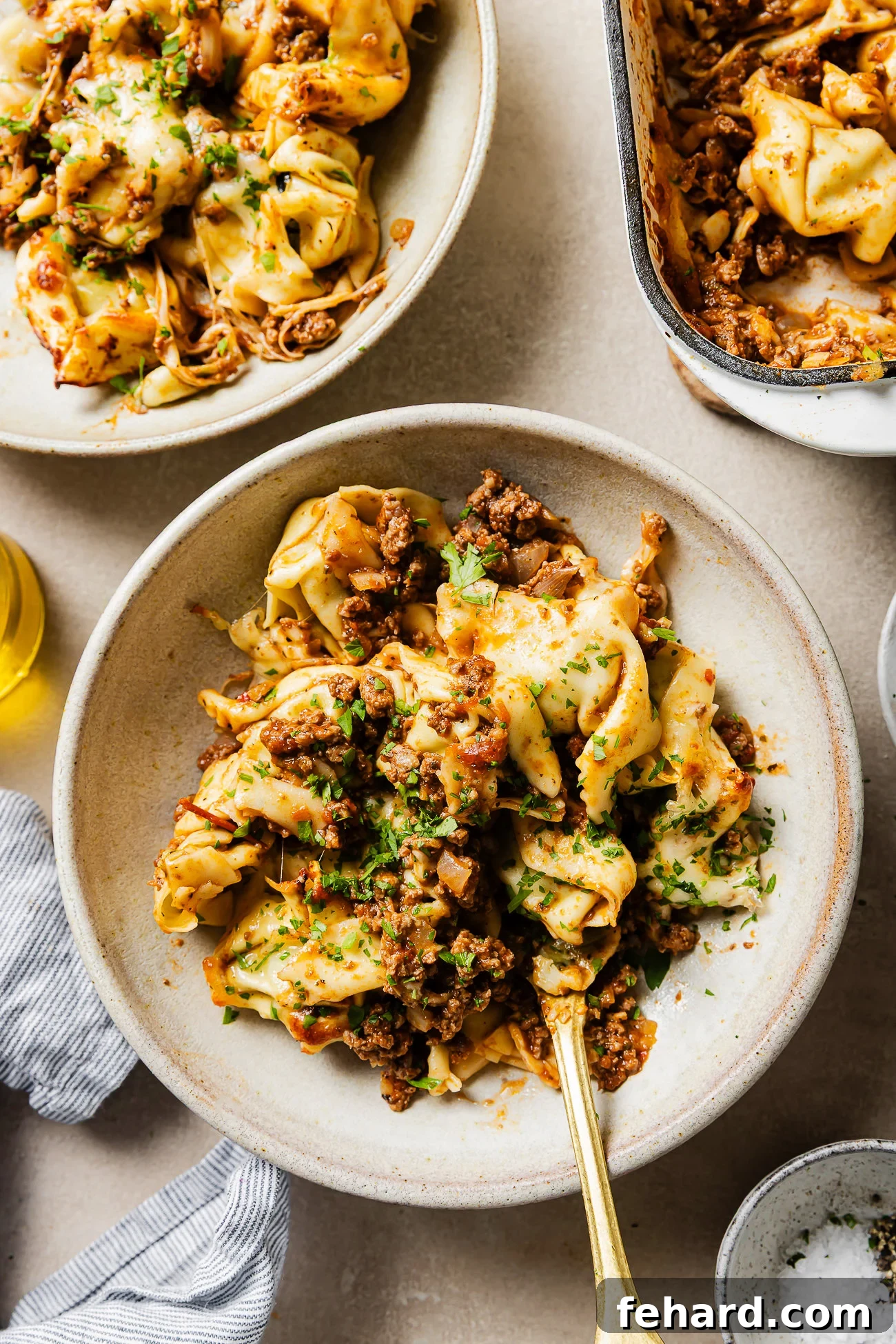 Cheesy Baked Tortellini with Savory Ground Beef Casserole 10 A bowl of tortellini pasta topped with a hearty meat sauce and fresh herbs, with a fork in the bowl. There are more servings of pasta in dishes nearby on a light-colored surface.
