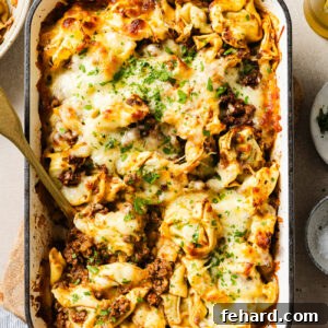 Cheesy Baked Tortellini with Savory Ground Beef Casserole 11 A baked pasta casserole in a white dish filled with cheese, ground meat, and herbs, topped with melted cheese and chopped parsley. A serving spoon is placed in the dish, ready for serving.