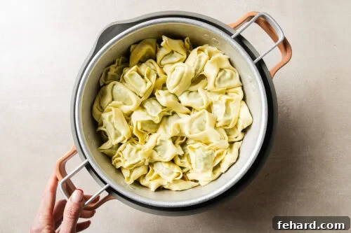 Cheesy Baked Tortellini with Savory Ground Beef Casserole 12 A hand holds a pot filled with uncooked tortellini pasta on a light-colored surface.