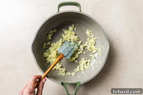 Cheesy Baked Tortellini with Savory Ground Beef Casserole 13 A hand stirs chopped onions in a large, light-colored pan with a green handle, using a spatula. The onions are being sautéed on a light-colored surface.