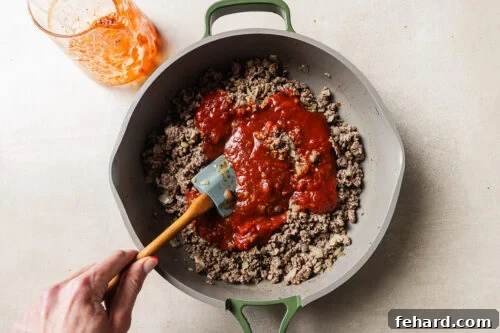 Cheesy Baked Tortellini with Savory Ground Beef Casserole 14 A hand stirs browned ground beef and chopped onions in a skillet with tomato sauce being mixed in. A glass with leftover sauce sits nearby on a light surface.