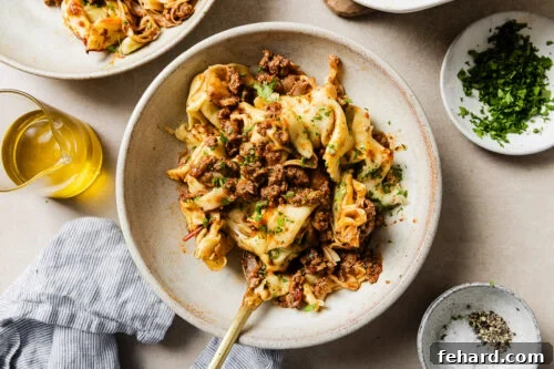 Cheesy Baked Tortellini with Savory Ground Beef Casserole 17 A bowl of pasta with crumbled ground meat, garnished with chopped herbs. Surrounding the bowl are a dish of salt and pepper, a small bowl of herbs, a glass container of olive oil, and a striped napkin.