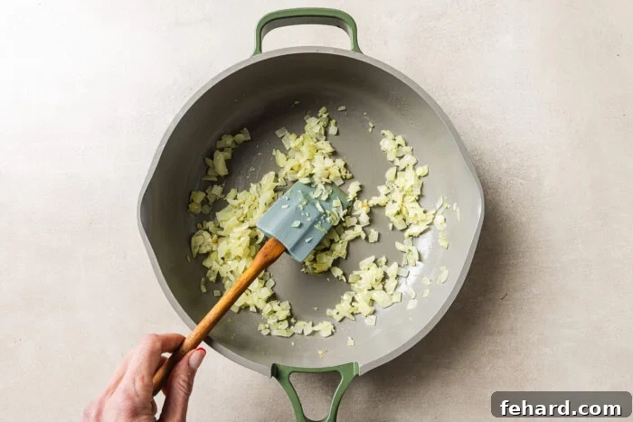 Cheesy Baked Tortellini with Savory Ground Beef Casserole 5 A hand stirs chopped onions in a large, light-colored pan with a green handle, using a spatula. The onions are being sautéed on a light-colored surface.
