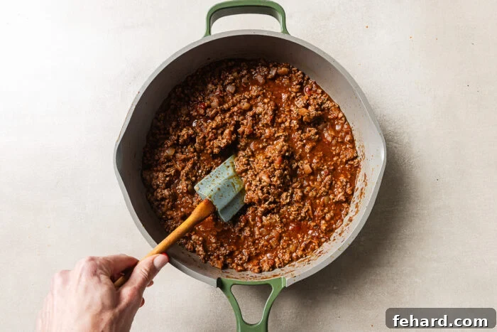 Cheesy Baked Tortellini with Savory Ground Beef Casserole 7 A hand stirs seasoned ground beef and tomato sauce with a green spatula in a large gray pot on a light countertop.