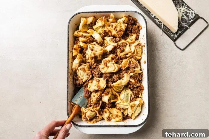 Cheesy Baked Tortellini with Savory Ground Beef Casserole 8 A hand holds a spatula in a baking dish filled with tortellini pasta and meat sauce. A block of Parmesan cheese and a grater sit on the surface nearby.