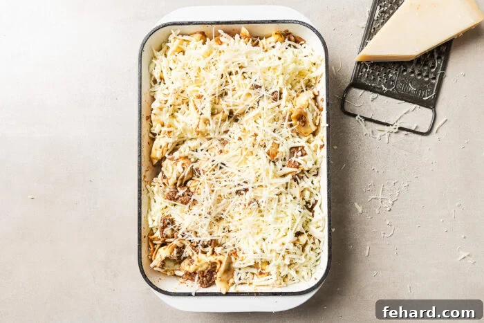 Cheesy Baked Tortellini with Savory Ground Beef Casserole 9 A rectangular baking dish filled with uncooked pasta topped with shredded cheese sits on a light surface. Next to it are a metal grater and a wedge of cheese, with cheese shavings scattered around.