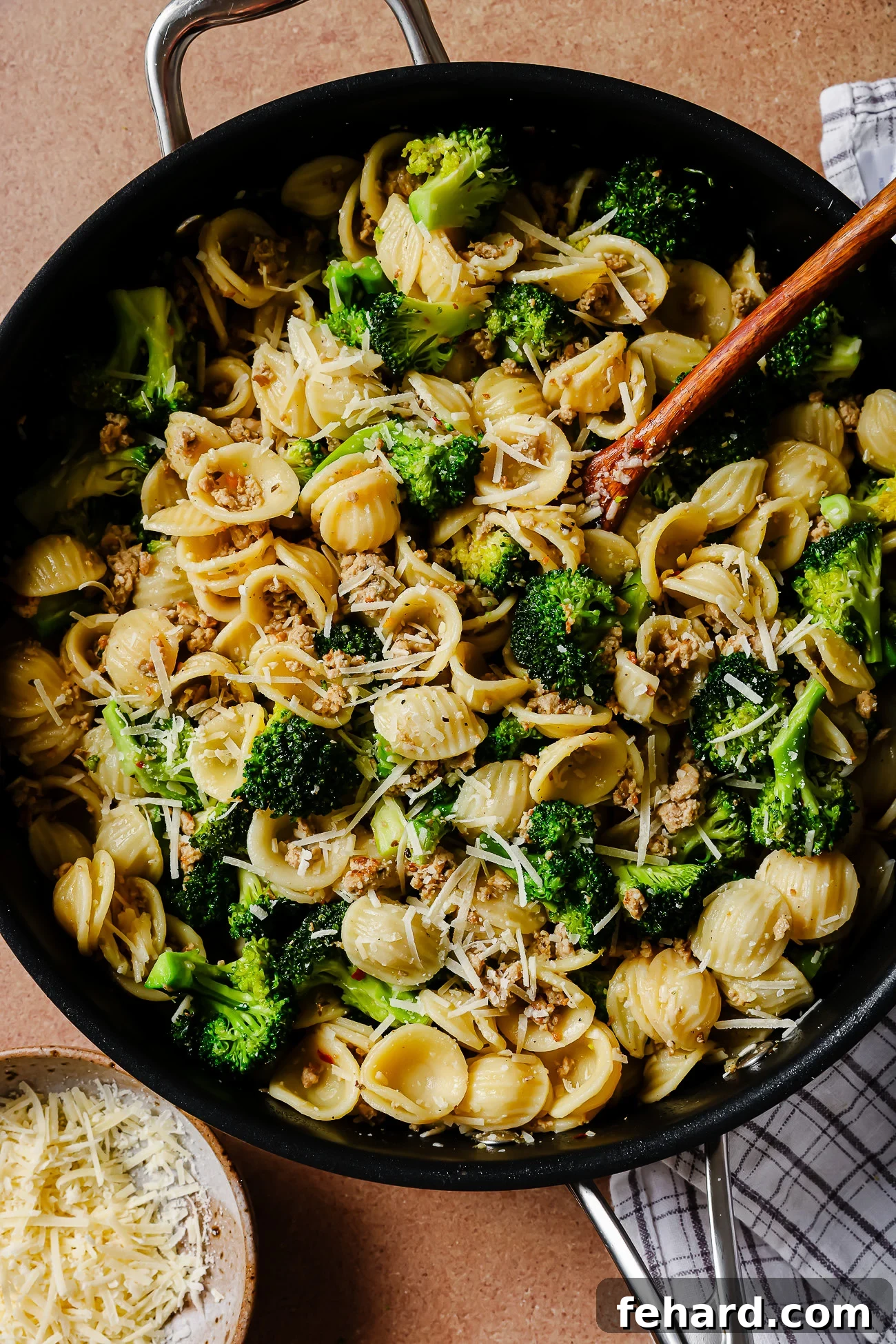 A skillet filled with orecchiette pasta, broccoli florets, crumbled sausage, and grated Parmesan cheese, with a wooden spoon and a bowl of extra cheese nearby on a striped cloth.