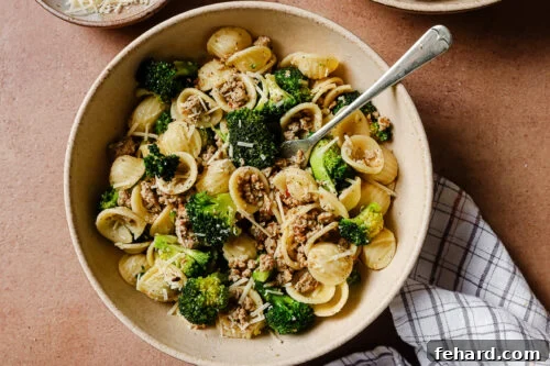 A bowl of orecchiette pasta mixed with cooked ground meat, broccoli florets, and grated cheese, with a fork in the bowl and a checked cloth napkin beside it.