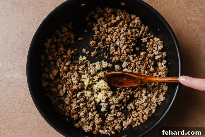 A hand stirs cooked ground meat and minced garlic in a black skillet with a wooden spoon on a brown countertop.