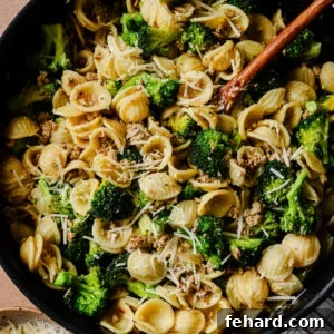 A skillet filled with orecchiette pasta, broccoli florets, crumbled sausage, and grated Parmesan cheese, with a wooden spoon and a bowl of extra cheese nearby on a striped cloth.