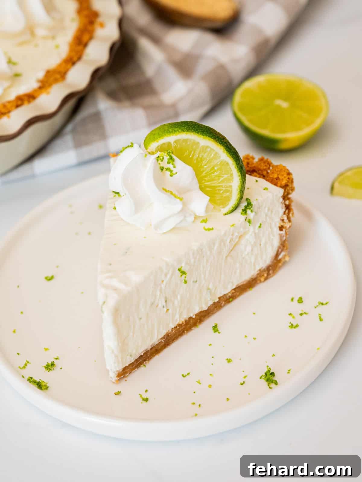 Easy Key Lime Pie Recipe Slice of homemade key lime pie with graham cracker crust on plate topped with piped whipped cream, a lime slice, and lime zest.