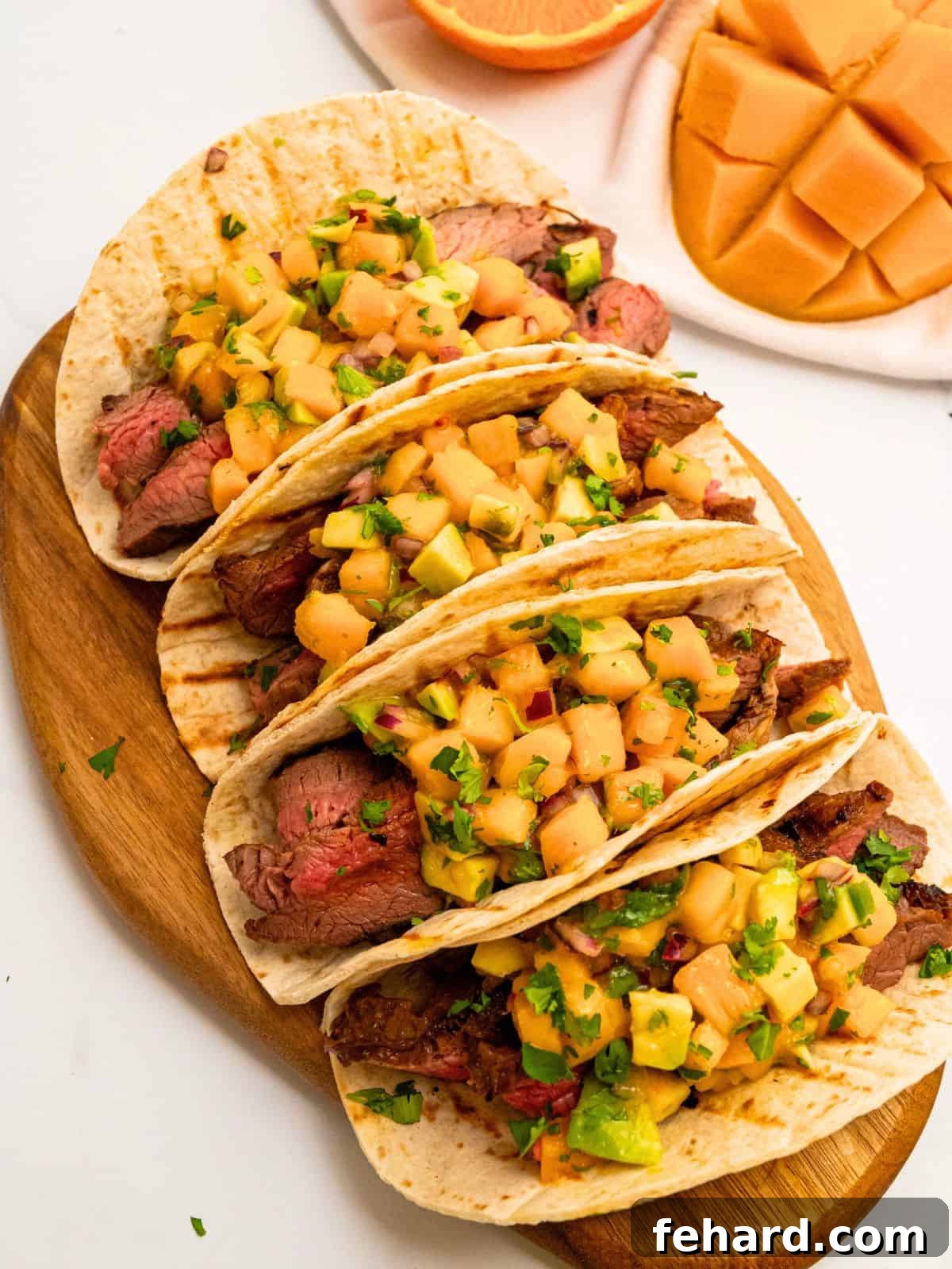 4 soft tortillas filled with sliced grilled marinated flank steak and topped with mango avocado salsa next to fresh mango.