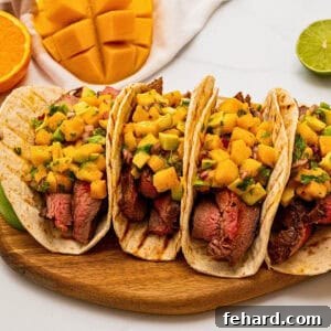 4 soft tortillas filled with sliced grilled marinated flank steak and topped with mango avocado salsa next to fresh mangoes, limes, and oranges.