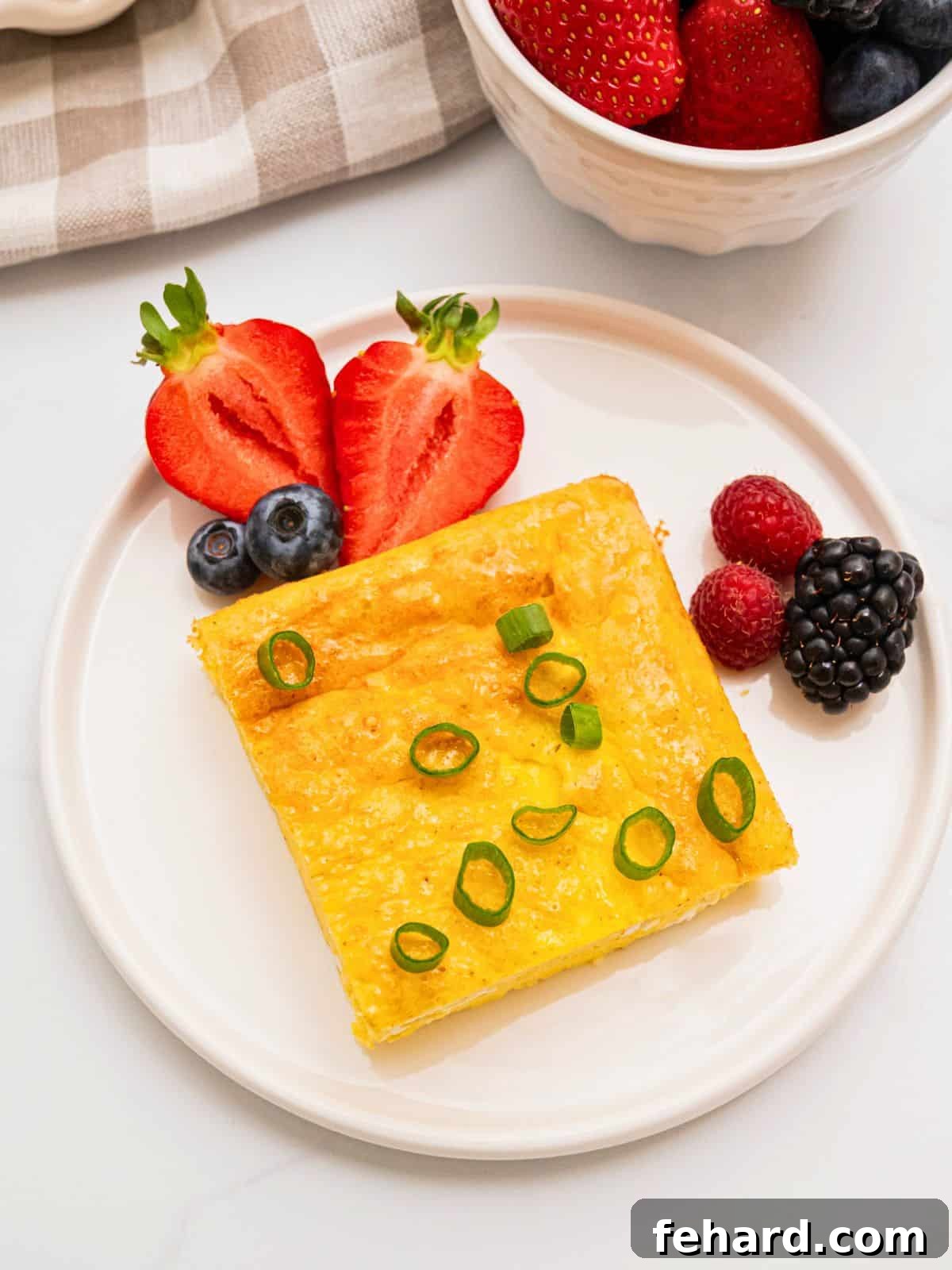 Baked Cottage Cheese Eggs Recipe Slice of cottage cheese egg bake on white plate topped with sliced green onions and berries on the side.