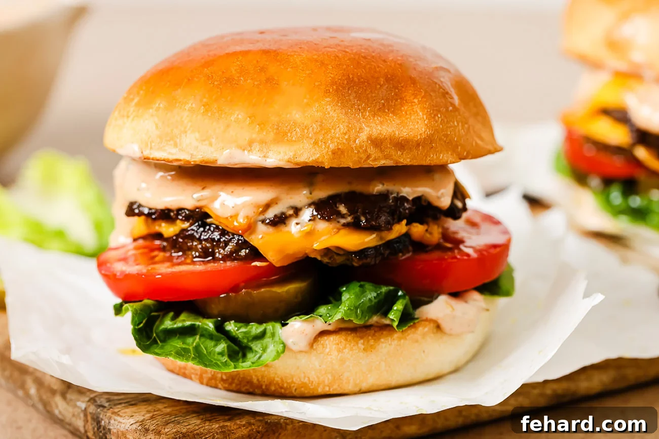 A close-up of a cheeseburger with two beef patties, melted cheddar cheese, lettuce, tomato slices, pickles, and sauce in a shiny, toasted bun, placed on parchment paper on a wooden surface.