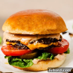 A close-up of a juicy cheeseburger with a golden bun, lettuce, tomato slices, melted cheese, two beef patties, and creamy sauce, served on parchment paper atop a wooden board.