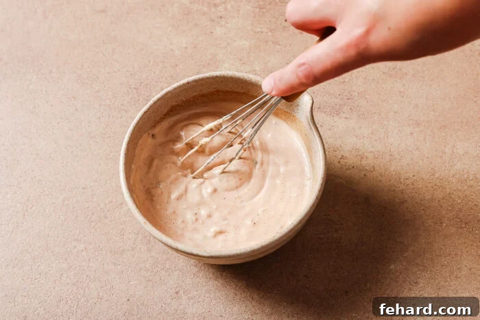 A hand whisking a creamy, light brown sauce or dressing in a beige ceramic bowl on a light brown surface.