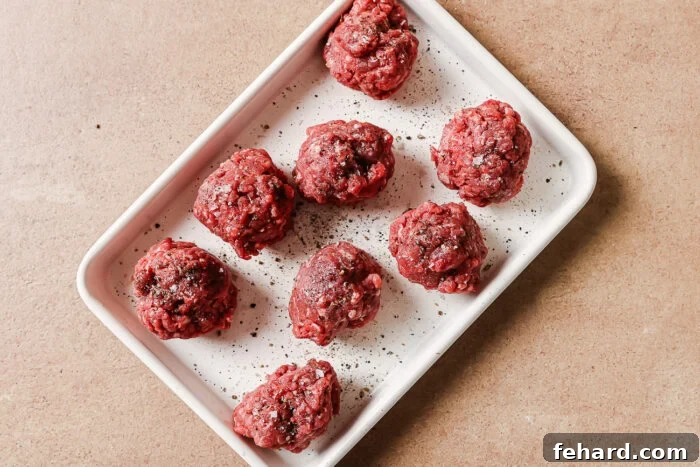 A white rectangular tray holds eight raw beef meatballs sprinkled with ground black pepper, set on a light brown surface.