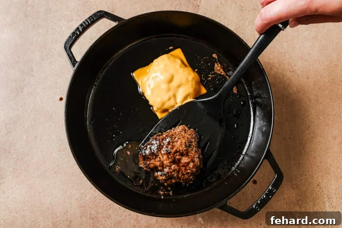 A hand uses a spatula to lift a cooked hamburger patty in a black skillet. Another patty with a slice of melted yellow cheese is also in the skillet on a beige countertop.