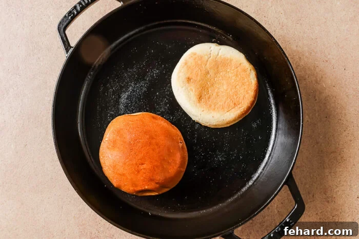 A cast iron skillet with two English muffins inside, one with a golden brown top and the other with a lighter, pale top, sitting on a beige surface.