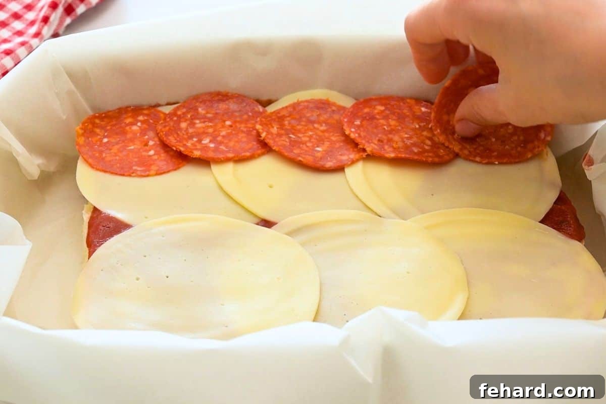 Layering pepperoni over bottom of slider rolls that have been topped with thin layer of pizza sauce and sliced provolone cheese.