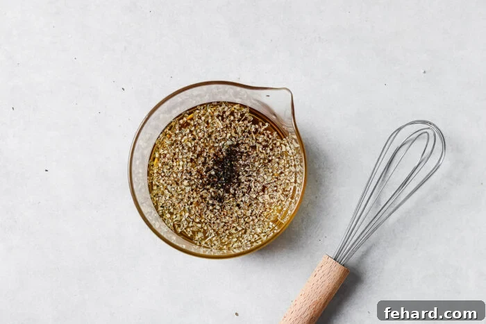 A glass measuring cup filled with oil and herbs sits on a light surface next to a metal whisk with a wooden handle.