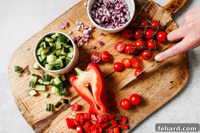 A hand slices cherry tomatoes on a wooden cutting board, surrounded by chopped cucumber, red bell pepper, diced red onion, and a bowl of whole and halved cherry tomatoes.