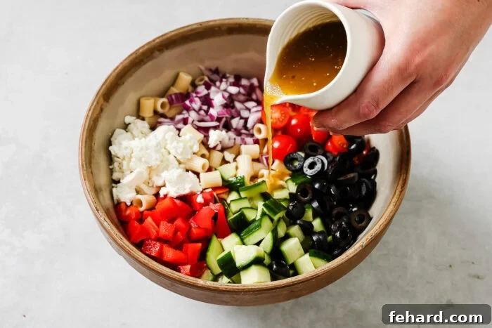 A hand pours dressing from a small white pitcher onto a bowl filled with chopped cucumbers, red bell peppers, cherry tomatoes, black olives, red onions, pasta, and crumbled feta cheese.