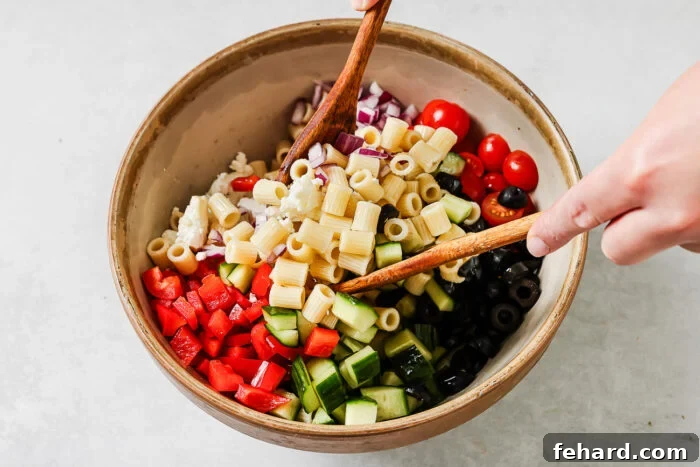 A person uses wooden utensils to mix a bowl filled with pasta, chopped red bell pepper, cucumber, cherry tomatoes, red onion, black olives, and feta cheese.
