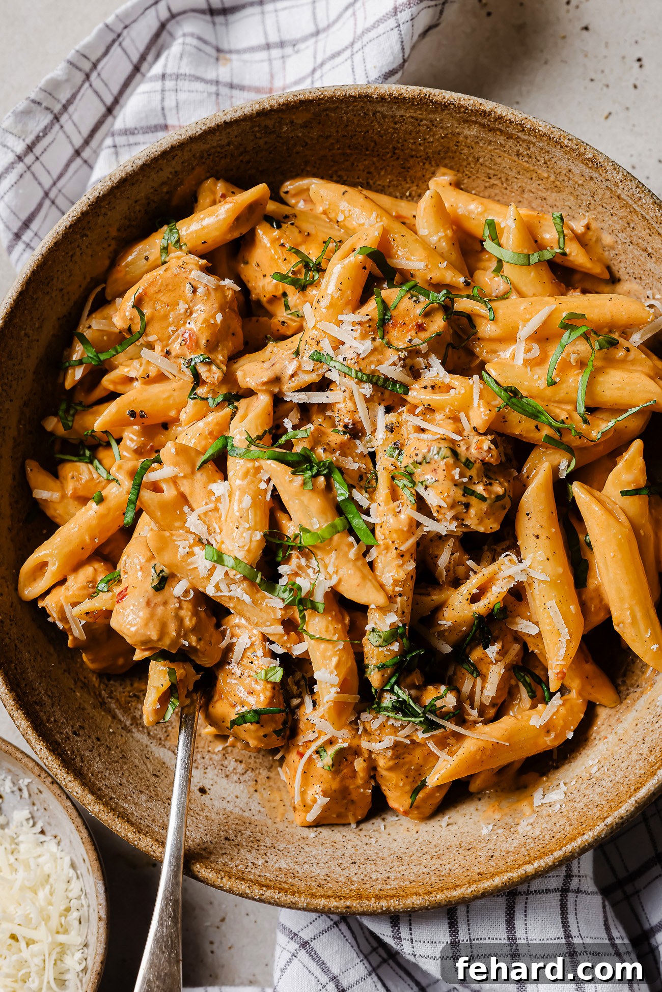 A bowl of creamy penne pasta with pieces of chicken, topped with grated cheese and chopped fresh herbs. A fork is placed in the bowl, and a checked cloth napkin is partially visible underneath.