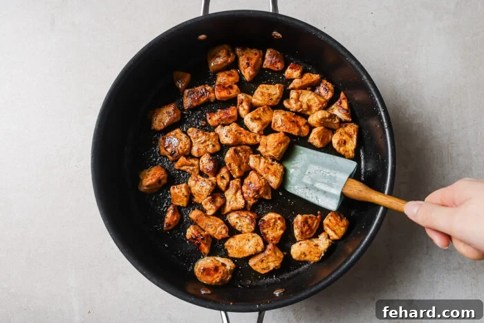 A hand holding a spatula stirs browned, seasoned chicken pieces in a black frying pan on a light-colored countertop.